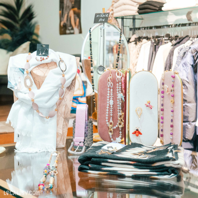 Un présentoir de bijoux et d'accessoires dans le magasin de vêtements pour femmes Dudilou à Hamoir, avec divers colliers, bracelets et boucles d'oreilles exposés sur des supports élégants. Il y a également des écharpes et des vêtements pliés en arrière-plan. Les bijoux sont variés, avec des perles, des pierres colorées et des pendentifs décoratifs.