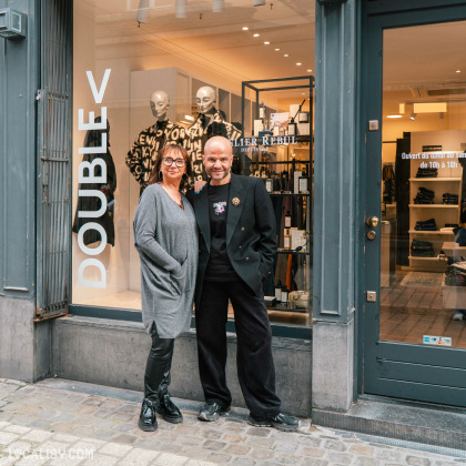 Devanture du magasin de vêtements pour femmes Double V à Liège, avec une vitrine qui expose des vêtements et une enseigne indique ATELIER REBUL DEPUIS 1895. Deux personnes debout devant le magasin. La porte affiche les horaires d'ouverture : Ouvert du lundi au samedi de 10h à 18h.