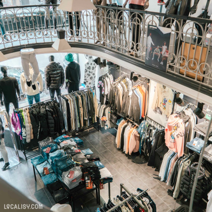 L'intérieur du magasin de vêtements Icon Store à Liège, avec des vêtements suspendus sur des cintres et des mannequins habillés, ainsi que des tables avec des piles de jeans. Le magasin est bien éclairé et présente une variété de vêtements, y compris des vestes, des sweats à capuche, des pantalons et des chemises. Il y a également un étage supérieur visible avec une balustrade en métal décoratif.