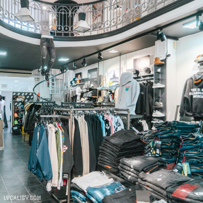 L'intérieur du magasin de vêtements Icon Store à Liège, avec des vêtements suspendus sur des cintres et empilés sur des étagères. On peut voir une variété de vêtements, y compris des vestes, des pantalons, des sweats à capuche et des jeans. Il y a également des accessoires comme des casquettes.