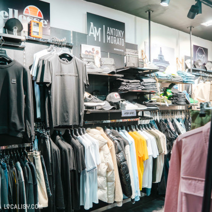 Des étagères et des portants remplis de vêtements, notamment des t-shirts, des vestes, des pulls et des pantalons dans le magasin de vêtements Icon Store à Liège. Les marques visibles incluent Antony Morato et Ellesse. Les vêtements sont bien organisés par type et couleur, et il y a également des accessoires comme des casquettes et des chaussures.