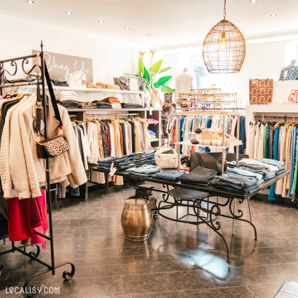 L'intérieur du magasin de vêtements pour femmes Chez Yo à Aywaille, avec des vêtements suspendus sur des cintres, des étagères remplies de vêtements pliés, et une table centrale avec des jeans et des accessoires. L'éclairage est chaleureux avec des spots au plafond et une grande lampe suspendue.