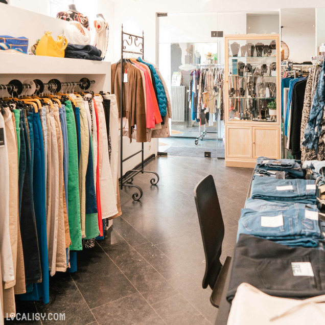 Des rangées de pantalons de différentes couleurs accrochés à des cintres sur la gauche, des vestes et manteaux suspendus sur un portant au centre, et des jeans pliés sur une table à droite dans le magasin de vêtements pour femmes Chez Yo à Aywaille. À l'arrière-plan, il y a une vitrine avec des accessoires et des bijoux, ainsi que d'autres vêtements suspendus.