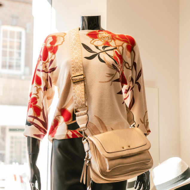 Un mannequin dans la vitrine du magasin de vêtements pour femmes Chez Yo à Aywaille. Le mannequin porte un pull à manches longues avec un motif floral rouge, beige et noir, et est accessoirisé avec un sac à bandoulière beige à deux poches.