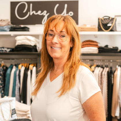 Une personne debout dans le magasin de vêtements pour femmes Chez Yo à Aywaille. En arrière-plan, on peut voir des étagères et des portants avec divers vêtements pliés et suspendus. Il y a également un sac à main noir avec une chaîne dorée sur une étagère. Le nom du magasin est visible sur un panneau au-dessus de la personne.