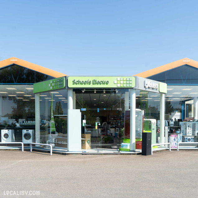 Devanture du magasin d'électroménager Schoofs Electro à Sprimont. Le magasin a une vitrine en verre avec des enseignes vertes et blanches. À l'intérieur, on peut voir divers appareils électroménagers exposés, tels que des machines à laver et des réfrigérateurs. Le bâtiment a un toit en pente avec des accents orange.