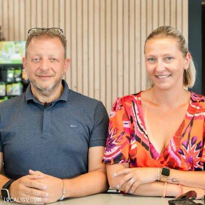 Dux personnes assises à une table dans un environnement intérieur au magasin d'électroménager Schoofs Electro à Sprimont. La personne à gauche porte un polo bleu foncé et des lunettes posées sur la tête. La personne à droite porte une robe colorée avec des motifs rouges, roses et noirs. Les deux personnes portent des montres intelligentes et des bracelets. En arrière-plan, il y a un mur en bois avec des lattes verticales et une étagère contenant des produits.