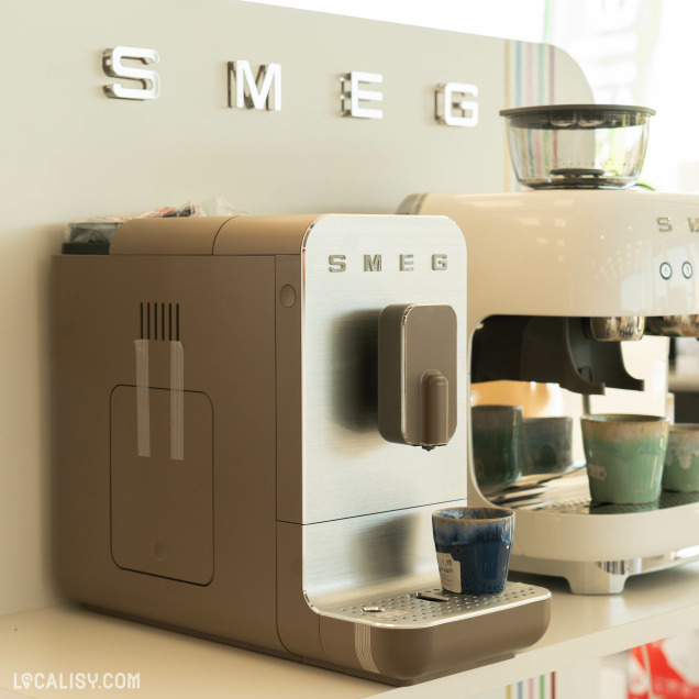 Deux machines à café de la marque SMEG exposées dans le magasin d'électroménager Schoofs Electro à Sprimont. La machine au premier plan est beige avec une façade en métal brossé, tandis que celle en arrière-plan est blanche. Des tasses en céramique colorées sont disposées en dessous des machines.