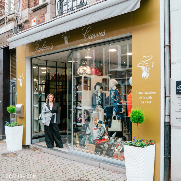 Devanture du magasin de vêtements pour femmes Fashion's Curves à Visé. La façade est dorée avec des inscriptions blanches indiquant le nom du magasin et un dessin stylisé d'une silhouette féminine. Une mannequin est placée à l'entrée, habillée de vêtements élégants. À l'intérieur, on peut voir des sacs à main et des vêtements exposés sur des mannequins.