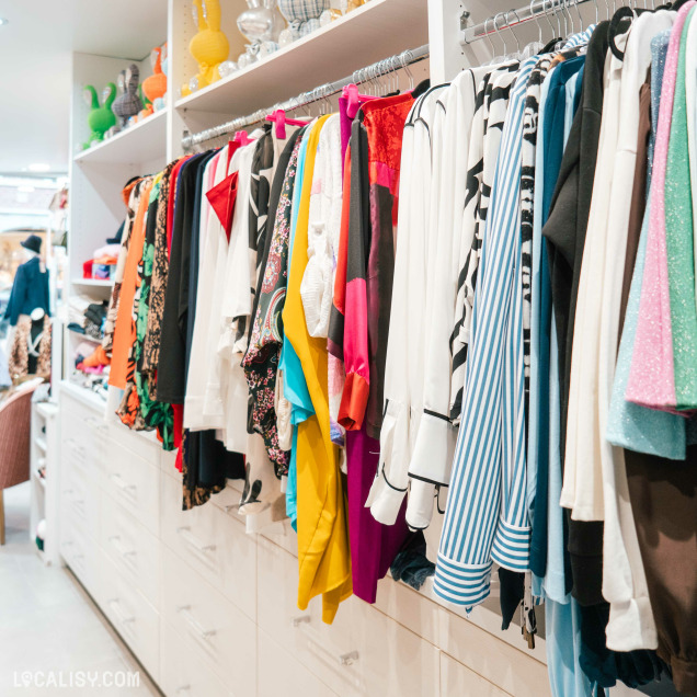 Une rangée de vêtements suspendus sur des cintres dans le magasin de vêtements pour femmes Fashion's Curves à Visé. Les vêtements sont de différentes couleurs et styles, allant des chemisiers aux robes. En dessous des vêtements, il y a des tiroirs blancs. Sur les étagères supérieures, il y a des objets décoratifs comme des vases et des sculptures colorées.