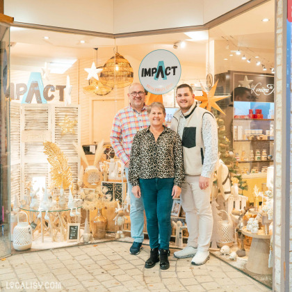 Devanture du magasin de décoration d’intérieur Impact Déco à Herve, avec trois personnes debout devant l'entrée. La vitrine du magasin est décorée avec divers objets de décoration, notamment des étoiles suspendues, des vases, des plantes séchées, et des figurines. Le logo du magasin est visible en haut de la vitrine avec le texte IMPACT et VOTRE ESPACE DECO.