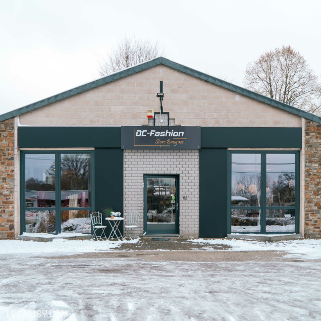 Devanture du magasin de vêtements pour femmes DC Fashion Store Baugnez à Malmedy. Le bâtiment a une structure en briques avec des accents de pierre sur les côtés. Il y a une enseigne noire avec le nom du magasin écrit en blanc. Devant le magasin, il y a une petite table ronde avec deux chaises, et le sol est couvert de neige.