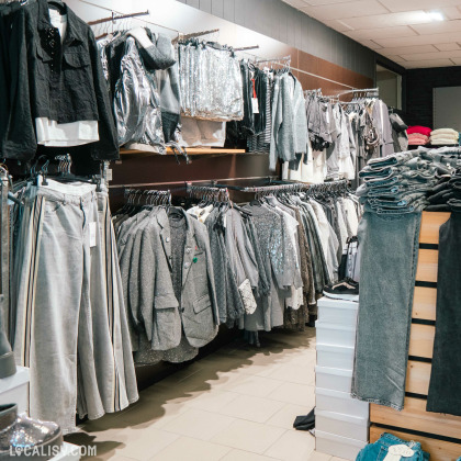 L'intérieur du magasin de vêtements pour femmes DC Fashion Store Baugnez à Malmedy, avec des vêtements principalement de couleur grise, accrochés sur des cintres et disposés sur des étagères. Il y a une variété de vêtements, y compris des vestes, des pantalons, des jupes, et des hauts, certains avec des paillettes et des tissus brillants.