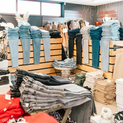 L'intérieur du magasin de vêtements pour femmes DC Fashion Store Baugnez à Malmedy, avec plusieurs piles de jeans de différentes couleurs et styles, soigneusement disposées sur des étagères en bois. Il y a également des vêtements suspendus à des cintres à l'arrière-plan, ainsi que des piles de pantalons beiges et verts sur le côté droit.