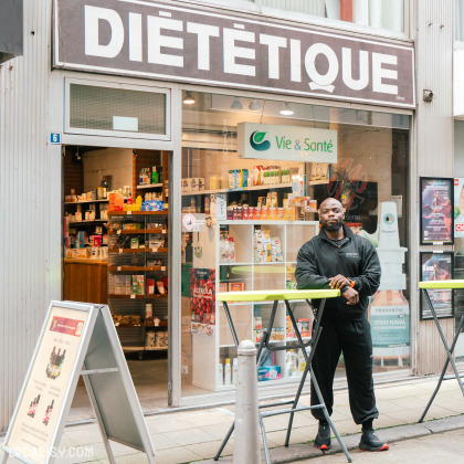 Devanture du magasin de nutrition sportive et diététique Vie et Santé à Liège. Le magasin a une grande enseigne avec le mot DIÉTÉTIQUE écrit en lettres blanches sur un fond marron. À l'intérieur, on peut voir des étagères remplies de divers produits diététiques et de suppléments nutritionnels. Devant le magasin, il y a un homme debout près de tables pliantes jaunes.