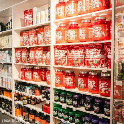 Des étagères remplies de divers produits de nutrition sportive, principalement des pots et des sachets de protéines en poudre de la marque 100% Whey Protein dans le magasin de nutrition sportive et diététique Vie et Santé à Liège. Les produits sont bien organisés et étiquetés, avec des prix visibles sur certaines étagères.