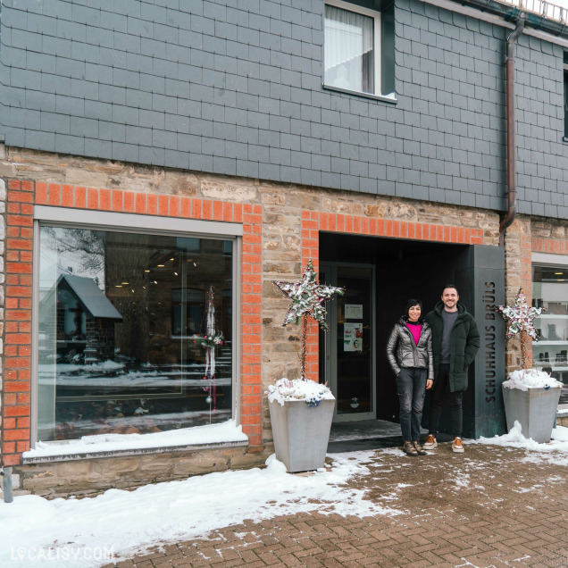 Devanture du magasin de chaussures Maison Brüls à Malmedy. Deux personnes se tiennent devant l'entrée du magasin, qui est décorée pour l'hiver avec des étoiles ornées de guirlandes et de neige. Le bâtiment est en pierre avec des accents de briques rouges autour des fenêtres et de la porte. Le sol est recouvert de neige, indiquant une saison hivernale.