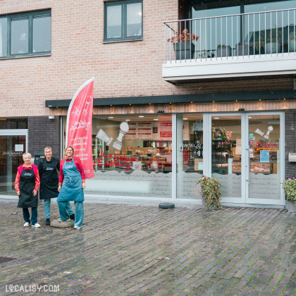 Devanture de la Sandwicherie et Traiteur Aux délices d'Embourg à Embourg. Devant l'établissement, trois personnes portant des tabliers noirs et bleus se tiennent debout. L'enseigne de la boutique est visible sur une grande vitre, avec un logo représentant un tampon et le nom de l'établissement écrit en rouge. Une bannière rouge à gauche de l'entrée affiche des informations supplémentaires sur les services offerts, tels que Pâté, Traiteur, Sandwiches, Épicerie, Charcuterie, Fromages.