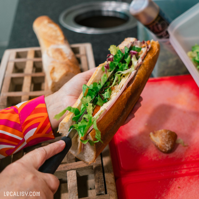 Une personne dans la Sandwicherie et Traiteur Aux délices d'Embourg à Embourg en train de préparer un sandwich avec une baguette de pain. La personne tient le sandwich d'une main et utilise un couteau de l'autre main pour ajuster le pain. Le sandwich est garni de légumes verts frais et de tranches de viande. En arrière-plan, on peut voir une baguette de pain sur une grille en bois et un plan de travail avec une planche à découper rouge.