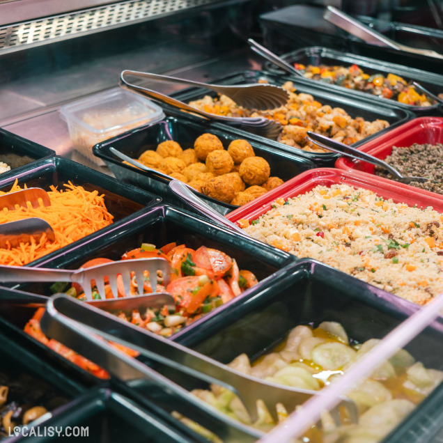 Un buffet de salades et de plats préparés dans la sandwicherie et traiteur Aux Délices d'Embourg à Embourg. On y voit plusieurs bacs contenant divers aliments tels que des carottes râpées, des boulettes, des salades de légumes, et du couscous. Chaque bac est équipé de pinces pour se servir.