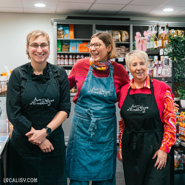 Trois personnes portant des tabliers noirs et bleus avec l'inscription Aux Délices d'Embourg. Elles se tiennent devant des étagères remplies de produits alimentaires variés, suggérant qu'elles travaillent dans la Sandwicherie et Traiteur Aux délices d'Embourg à Embourg.