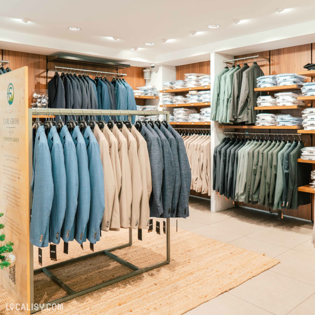 L'intérieur du magasin de vêtements pour hommes Thimister Men à Herve, avec des étagères et des portants remplis de vestes et de chemises soigneusement organisées. Les vestes sont de différentes couleurs, principalement des tons de bleu, beige et gris. Les chemises sont pliées et empilées sur les étagères.