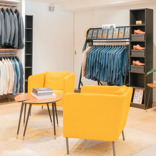 Des costumes et des vestes suspendus sur des portants, avec une variété de couleurs allant du bleu au beige dans le magasin de vêtements pour hommes Thimister Men à Herve. Il y a également des chaussures en cuir marron exposées sur des étagères. Au centre de la pièce, il y a deux fauteuils jaunes vifs et une petite table ronde avec des livres posés dessus.