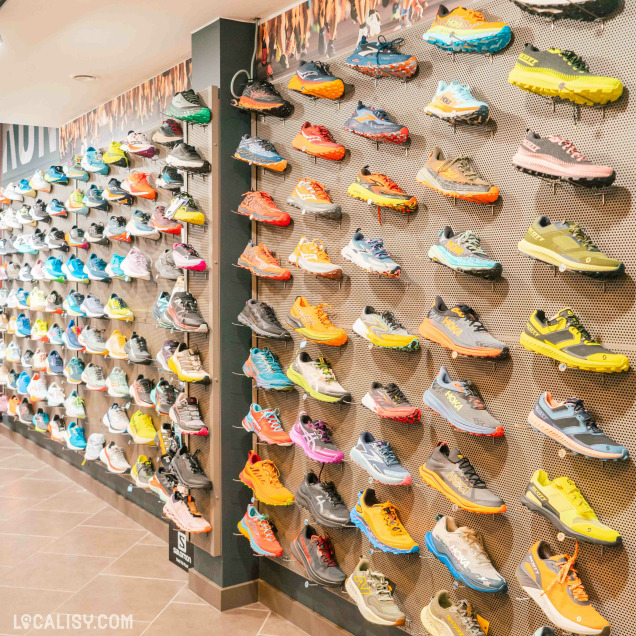 Un mur du magasin d'articles de course à pied Xrun à Malmedy, avec une large sélection de chaussures de course exposées. Les chaussures sont disposées en rangées verticales et horizontales, avec une grande variété de couleurs et de modèles. Le mur de gauche présente des chaussures principalement de couleur claire, tandis que le mur de droite présente des chaussures de couleur plus vive et variée.