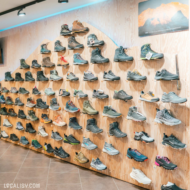 Un mur d'exposition recouvert de nombreuses paires de chaussures de randonnée et de course, disposées de manière ordonnée sur des étagères en bois dans le magasin d'articles de course à pied Xrun à Malmedy.. Chaque paire de chaussures est soigneusement placée pour être bien visible. En haut à droite, il y a une photo d'un paysage montagneux.