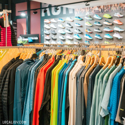 L'intérieur du magasin d'articles de course à pied Xrun à Malmedy. On y voit un présentoir avec une variété de vêtements de sport colorés suspendus sur des cintres. En arrière-plan, il y a un mur avec de nombreuses paires de chaussures de course exposées.