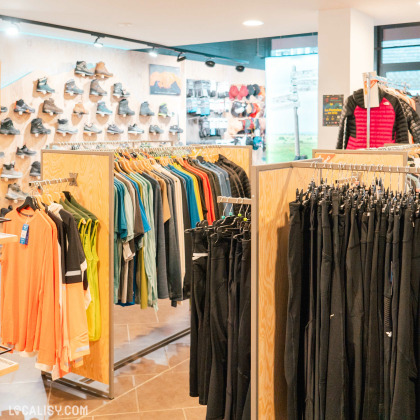 L'intérieur du magasin d'articles de course à pied Xrun à Malmedy, avec des étagères et des présentoirs remplis de vêtements de sport, notamment des t-shirts, des pantalons et des vestes. À l'arrière-plan, il y a un mur avec des chaussures de course exposées.