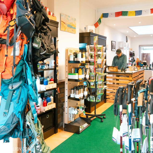 L'intérieur du magasin de matériel de camping Kmino-Outdoor à Esneux, avec divers équipements de camping tels que des sacs à dos, des bouteilles d'eau, des bâtons de marche et d'autres accessoires de plein air. Un homme se tient derrière un comptoir en bois, probablement en train de travailler ou d'aider un client. Le magasin est bien organisé avec des étagères et des présentoirs remplis de produits.