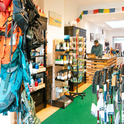 L'intérieur du magasin de matériel de camping Kmino-Outdoor à Esneux, avec divers équipements de camping tels que des sacs à dos, des bouteilles d'eau, des bâtons de marche et d'autres accessoires de plein air. Un homme se tient derrière un comptoir en bois, probablement en train de travailler ou d'aider un client. Le magasin est bien organisé avec des étagères et des présentoirs remplis de produits.