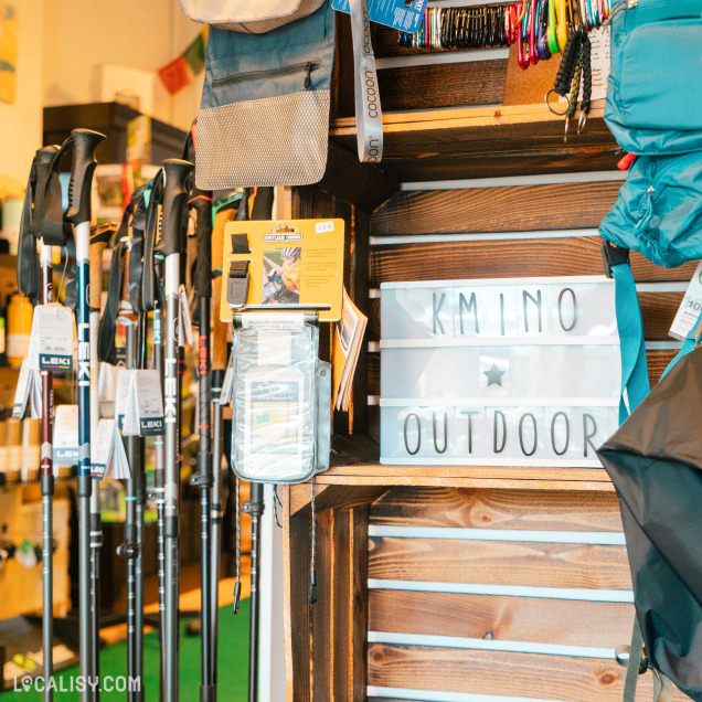 L'intérieur du magasin de matériel de camping Kmino-Outdoor à Esneux, avec divers équipements de camping tels que des sacs de couchage suspendus, des sacs à dos, des tentes et d'autres accessoires de camping. Une plante en pot est placée sur une table en bois au centre de l'image, entourée de brochures et de dépliants. L'agencement du magasin est bien organisé, avec des étagères en bois contenant les articles de camping.