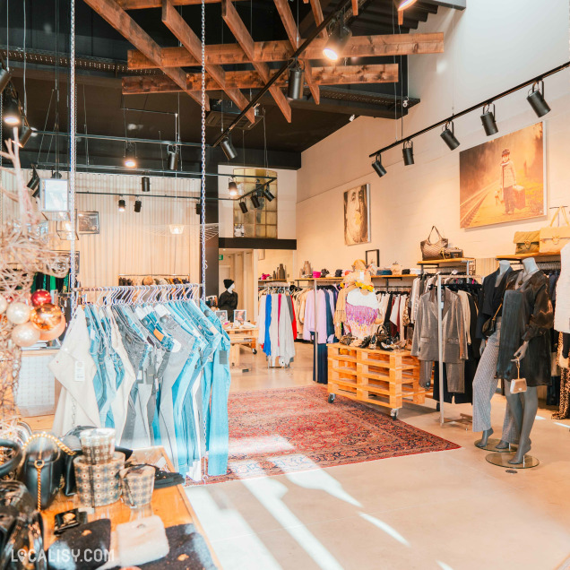 L'intérieur du magasin de vêtements pour femmes Ma.Dam à Herve, avec des poutres en bois apparentes au plafond et un éclairage suspendu. Il y a plusieurs portants de vêtements avec des jeans, des vestes, des robes et d'autres articles de mode. On peut également voir des mannequins habillés, des sacs à main exposés et un tapis rouge au sol.