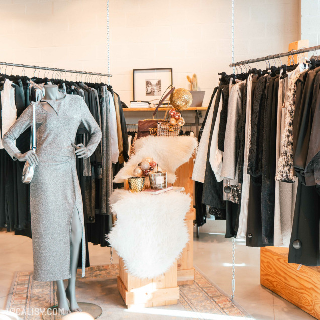 L'intérieur du magasin de vêtements pour femmes Ma.Dam à Herve. On y voit un mannequin portant une robe argentée à manches longues avec une fente sur le côté. Autour du mannequin, il y a des portants de vêtements avec divers articles de mode, principalement dans des tons neutres comme le noir, le gris et le blanc. Au centre, une table décorée avec des objets tels que des boules dorées et argentées, des bougies et des tissus en fausse fourrure blanche.