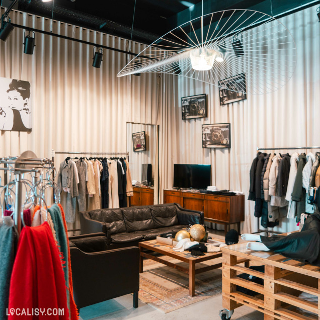 L'intérieur du magasin de vêtements pour femmes Ma.Dam à Herve, avec des portants de vêtements avec divers articles suspendus, des manteaux et des robes. Il y a également un canapé en cuir noir et une table basse en bois avec des décorations dorées. Des étagères en bois et une télévision sont visibles à l'arrière-plan. Le magasin est décoré avec des photos en noir et blanc accrochées aux murs et un grand luminaire moderne suspendu au plafond.