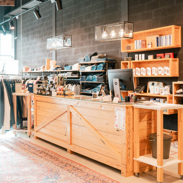 L'intérieur du magasin de vêtements pour femmes Ma.Dam à Herve. On y voit un comptoir en bois clair avec un ordinateur HP, des fournitures de bureau et divers articles exposés. Derrière le comptoir, il y a des étagères avec des vêtements pliés, principalement des jeans et des pulls. À gauche, un portant avec des vêtements suspendus est visible.