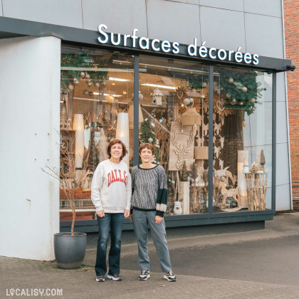 Devanture du magasin d'ameublement et de décoration Surfaces Décorées à Battice. La vitrine du magasin est décorée avec divers objets de décoration, notamment des lampes, des ornements et des décorations de Noël. Deux personnes se tiennent debout devant le magasin.