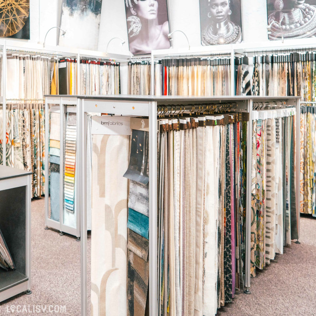 L'intérieur du magasin d'ameublement et de décoration appelé Surfaces Décorées à Battice, avec plusieurs étagères et présentoirs remplis de tissus de différentes couleurs et motifs. Les tissus sont suspendus sur des cintres, permettant aux clients de les examiner facilement. Des panneaux décoratifs sont accrochés au mur en arrière-plan, ajoutant une touche esthétique à l'espace.