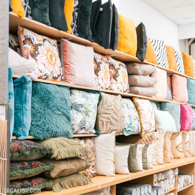 Une étagère dans le magasin d'ameublement et de décoration Surfaces Décorées à Battice, remplie de coussins de différentes tailles, couleurs et motifs. Les coussins sont disposés en plusieurs rangées, avec des motifs floraux, des textures en peluche, des couleurs unies et des motifs géométriques.