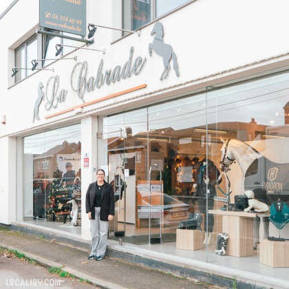 Devanture du magasin de matériel d'équitation Sellerie La Cabrade à Retinne, avec une grande vitrine en verre qui permet de voir l'intérieur, où sont exposés divers équipements équestres, y compris des selles, des tapis de selle et des mannequins de chevaux. Le nom du magasin est affiché en grandes lettres cursives au-dessus de la vitrine, accompagnées de silhouettes de chevaux.