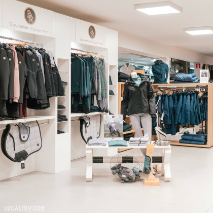 L'intérieur du magasin de matériel d'équitation Sellerie La Cabrade à Retinne, avec des étagères et des portants remplis de vêtements d'équitation, tels que des vestes, des manteaux et des pantalons. Il y a également des équipements pour chevaux, comme des tapis de selle. Un mannequin porte une tenue d'équitation complète. Une table au centre présente divers articles, et un panier en métal contient des accessoires.