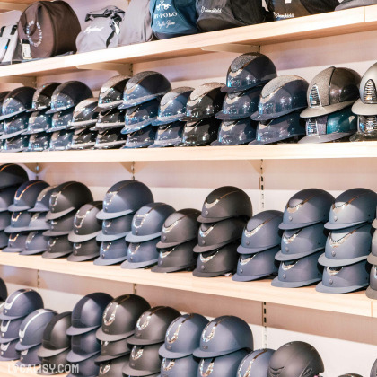 Plusieurs étagères remplies de casques d'équitation dans le magasin de matériel d'équitation Sellerie La Cabrade à Retinne. Les casques sont soigneusement disposés en rangées, avec différents styles et designs visibles. Certains casques ont une finition brillante, tandis que d'autres ont une finition mate. Il y a aussi des sacs et d'autres accessoires sur l'étagère du haut.