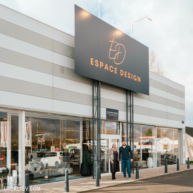 Devanture du magasin de luminaires Espace Design à Sprimont. Le bâtiment est moderne avec une grande enseigne noire portant le logo et le nom Espace Design en lettres dorées. La vitrine est en verre, permettant de voir l'intérieur du magasin où sont exposés divers articles de décoration et de design. Deux personnes sont visibles à l'entrée du magasin.