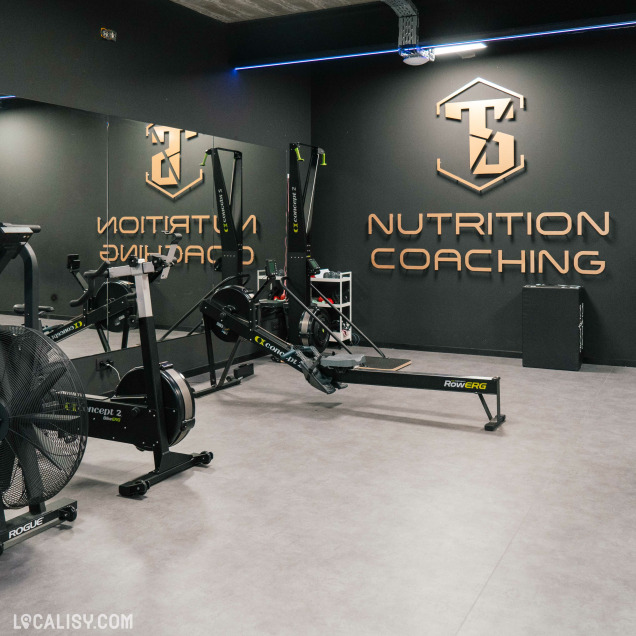 L'intérieur du magasin de coaching nutritionnel TS Nutrition Coaching à Ans. On y voit une salle de sport moderne avec plusieurs équipements de fitness, notamment des rameurs Concept2 et un vélo d'exercice Rogue. La salle est bien éclairée et propre, avec un sol en carrelage gris. Il y a également un grand miroir sur le mur de gauche, reflétant les équipements de fitness.