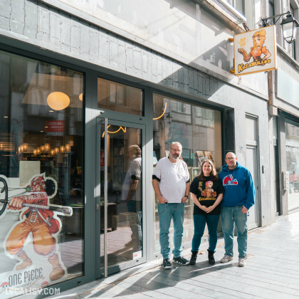 Devanture de la librairie de bandes dessinées Kazabulles à Liège. Trois personnes se tiennent devant l'entrée du magasin, qui a une grande vitrine en verre. À gauche de l'entrée, il y a une grande affiche de l'anime One Piece représentant un personnage avec une lance. Au-dessus de la porte, une enseigne avec le logo et le nom Kazabulles est visible. L'intérieur du magasin semble bien éclairé avec des lumières suspendues et des étagères visibles à travers la vitrine.