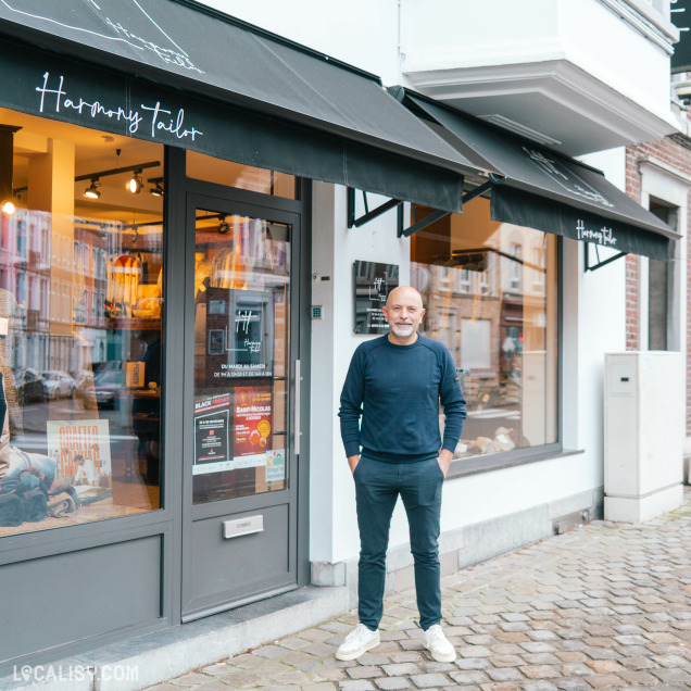 Devanture du magasin de vêtements pour hommes Harmony Tailor à Verviers. Un homme se tient devant l'entrée du magasin, les mains dans les poches. Le magasin a une enseigne noire avec le nom Harmony Tailor écrit en blanc. À travers la vitrine, on peut voir des vêtements et des accessoires exposés à l'intérieur. Il y a également plusieurs affiches et autocollants sur la porte vitrée, dont une mentionnant les horaires d'ouverture et une autre annonçant des promotions.