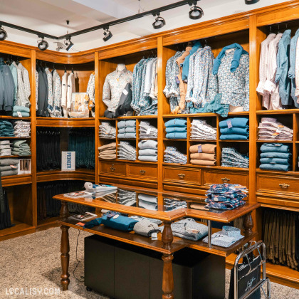 Des étagères en bois remplies de chemises, de pulls et de pantalons soigneusement pliés et disposés dans le magasin de vêtements pour hommes Harmony Tailor à Verviers. Il y a une table en bois au centre avec des vêtements pliés dessus. L'éclairage est fourni par des spots au plafond, mettant en valeur les vêtements exposés.