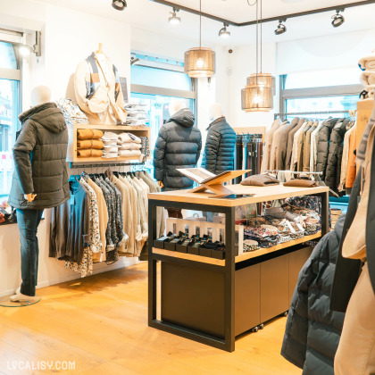 Plusieurs mannequins habillés en vêtements d'hiver, des étagères et des portants remplis de vêtements dans le magasin de vêtements pour hommes Harmony Tailor à Verviers. Au centre, il y a une table d'affichage avec des accessoires comme des ceintures et des montres. Le magasin est bien éclairé et présente une variété de vêtements d'hiver, notamment des manteaux, des vestes et des pulls.